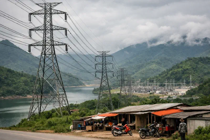 High-voltage transmission towers crossing northern Laos, with a hydro landscape behind them, capturing the shift from dam-building to corridor-based regional power trade.