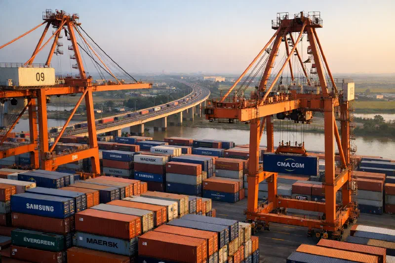 Aerial view of Haiphong port with container cranes and stacked shipping containers, with expressway visible in background connecting to industrial zones